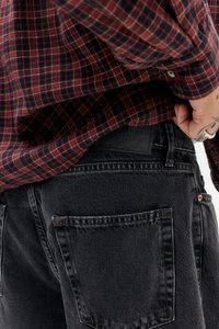 Moški v rdeči in črni karirasti srajci jo dviga, da pokaže zadnji žep črnih kavbojk, na roki so vidni tetovaže.