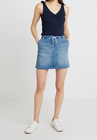 Jeansrock in Hellblau mit strukturierten Paneelen, lockerer Passform und Kordelzugbund. Kombiniert mit einem navyblauen Tanktop und weißen Turnschuhen.