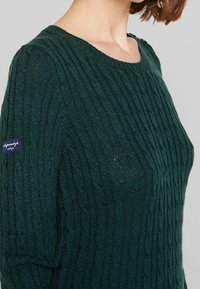 Zielony sweter wykonany na drutach z żebrowaną fakturą, okrągłym dekoltem i małą naszywką z logo na lewym ramieniu. Szczegółowy wzór w warkocze.