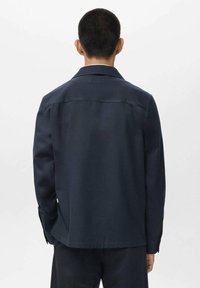 Camisa de manga larga color marino con un cuello extendido, textura suave y detalles de costura mínimos, mostrada desde la parte posterior sobre un fondo blanco.