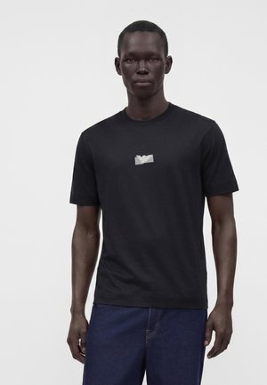 Homme portant un t-shirt noir à manches courtes avec un petit logo blanc sur la poitrine et un pantalon bleu foncé, debout devant un fond uni.