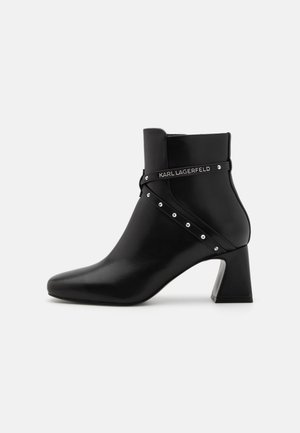 Black leather ankle boot with a pointed toe and block heel. Features a decorative strap with silver studs and a logo detail. Smooth finish.