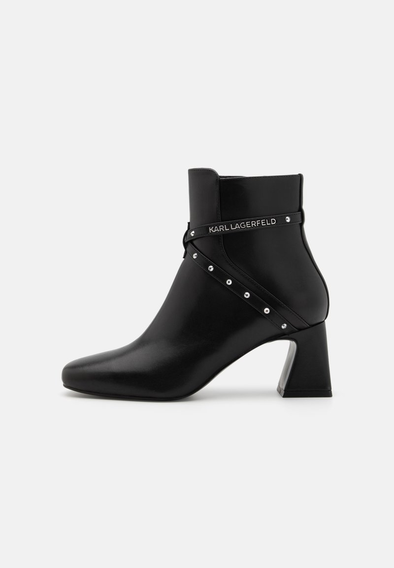 Black leather ankle boot with a pointed toe and block heel. Features a decorative strap with silver studs and a logo detail. Smooth finish.