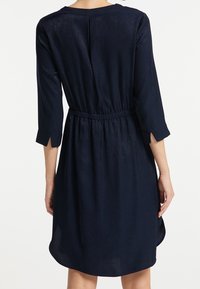 Vestido azul marino con cintura fruncida, mangas tres cuartos, un escote en la espalda con forma de llave y puños divididos. Tejido suave con un ligero brillo.