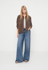 Brązowa koszula sztruksowa z przednimi kieszeniami, w połączeniu z jasnoniebieskimi, szerokimi spodniami dżinsowymi. Model stoi na tle jednolitego białego tła.