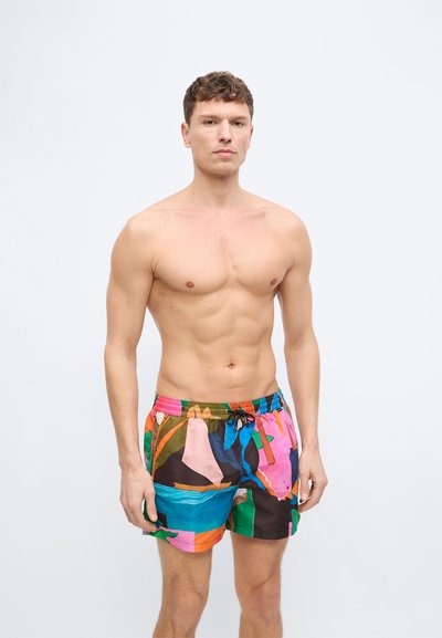 Άνδρας χωρίς πουκάμισο με κοντά καφέ μαλλιά, φορώντας πολύχρωμα μαγιό με αφηρημένο μοτίβο, στέκεται μπροστά σε έναν απλό λευκό φόντο.