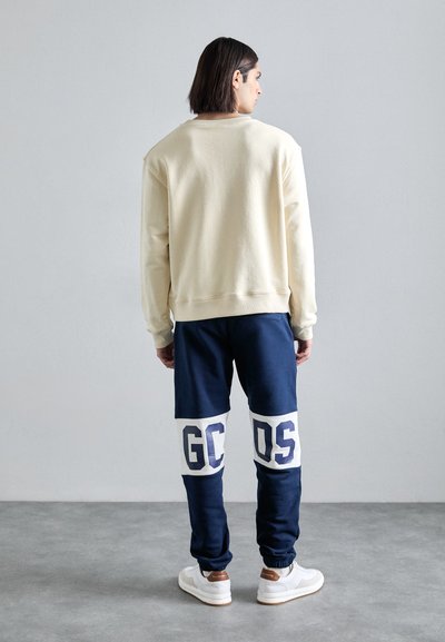 Sweatshirt crème avec des poignets côtelés, associé à un pantalon de jogging bleu marine arborant de grands imprimés blancs "GC" et "DS" sur les jambes inférieures, complété par des baskets blanches.