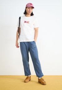 T-shirt crème avec un graphisme coloré, un pantalon de survêtement bleu et des chaussures beiges avec des lacets jaunes. Une casquette rose clair complète la tenue.