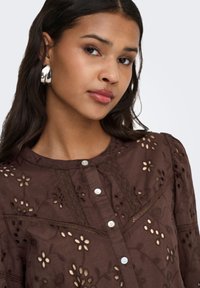 Braune bestickte Bluse mit floralen Cutouts, mit rundem Ausschnitt, Knopfleiste und strukturierten Spitzenakzenten. Sichtbare silberne Creolen.