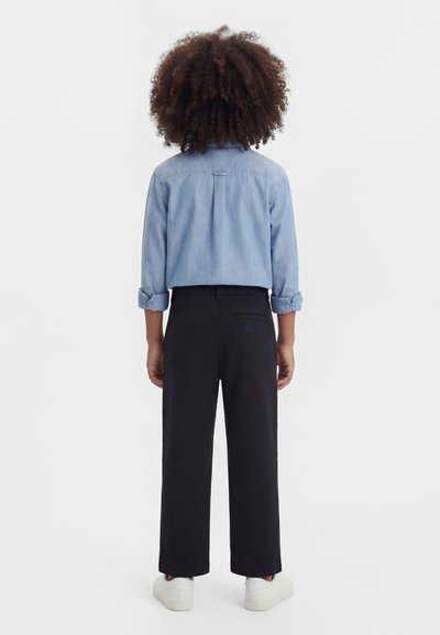 Persoană tânără cu păr creț și închis la culoare, purtând o cămașă din denim de culoare albastru deschis, pantaloni negri și adidași albi, stând cu fața în direcția opusă.