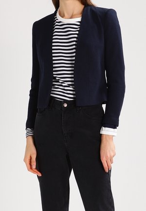 Vrouw draagt een navyblauwe blazer over een zwart-wit gestreept shirt met lange mouwen, gecombineerd met zwarte high-waisted jeans.