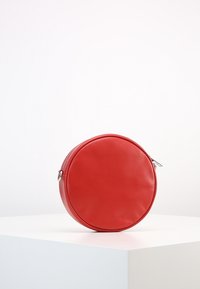 Röd cirkulär väska gjord av slät läder. Har en dragkedjestängning och präglande varumärkeslogotyp på framsidan. Enfärgad yta utan mönster.