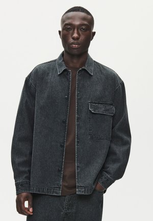 Un homme se tient devant un fond blanc uni, vêtu d’une veste en jean foncé boutonnée avec une poche poitrine et d’un pantalon assorti.