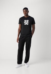 Svart T-skjorte med hvitt grafisk design på forsiden, kombinert med svarte bukser og hvite sneakers, med en avslappet passform og glatt tekstur.