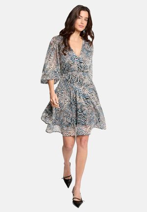 Femme marchant, portant une robe fluide mi-longue à motifs plume bleu et beige, et des escarpins noirs à bout pointu.