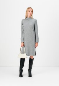 Siva obleka z dolgimi rokavi in visokim ovratnikom, z bočnima razporkoma, v kombinaciji z črnimi škornji do kolen in majhnim belim ročnim torbico. Gladka tekstura.