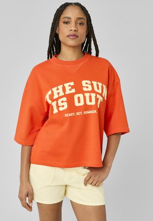 Jeune femme avec de longues tresses portant un t-shirt orange surdimensionné avec l'inscription "LE SOLEIL EST DEHORS" et un short beige clair.