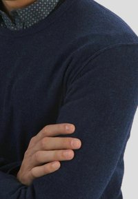 Donkerblauwe zachte gebreide trui met een ronde hals en een gestructureerde stof, gedragen over een patternd shirt. Handen gekruist over de arm.