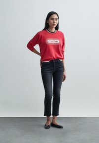 Sarkans džemperis ar baltu uzrakstu "mother", melni akcenti, kopā ar tumšām slaidām džinsa biksēm un melnām dzīvoklām kurpēm, ko valkā stāvoša figūra.