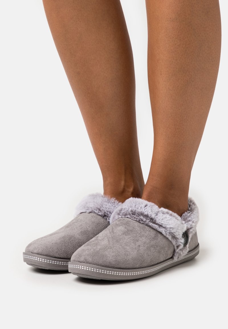 Jambes portant des chaussons gris sans lacets avec doublure en fourrure moelleuse et détail bouton, sur fond blanc.