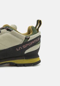 Zelená a béžová semišová lezečka s černou gumovou podrážkou, žlutými akcenty a růžovým logem. Obsahuje zesílenou patu a strukturovaný jazyk.