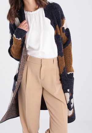 Frau trägt beige Hose, weiße Bluse und lange, gemusterte Strickjacke in Marineblau, Braun und Cremefarben.