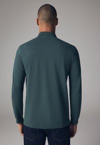 Langærmet polo shirt i mørk teal, lavet af blødt stof, med krave og enkel kant, parret med mørk denim jeans.