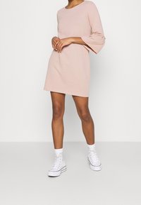 Ljusrosa klänning med rundad halsringning och trekvarts ärmar, tillverkad av mjukt tyg; kombinerad med vita hög-top sneakers.