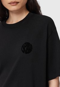 T-shirt di cotone nero con un logo rotondo e testurizzato in tonalità scura sul lato sinistro del petto. Maniche corte e scollo a girocollo.