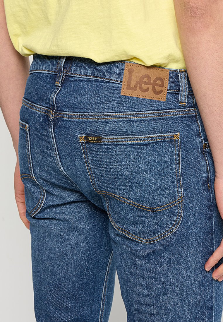 Niebieskie dżinsy w prostym kroju, z żółtym przeszyciem, tylnymi kieszeniami oraz brązową skórzaną łatą z logo na pasku.