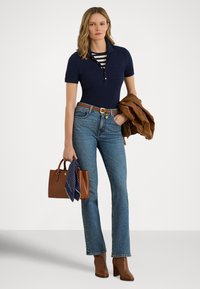 Μπλε πόλο μπλούζα με γιακά με κουμπιά, φορεμένη πάνω από ριγέ πουκάμισο, συνδυασμένη με ανοιχτό μπλε τζιν καμπάνα, καφέ ζώνη, μπότες με τακούνι και καφέ τσάντα χειρός.