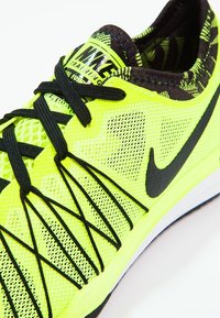 Neon žlutá tréninková bota Nike s černými tkaničkami, černým logem swoosh a vzorovanou vnitřní podšívkou, nakloněná tak, aby ukázala detaily svrchní části a strany.