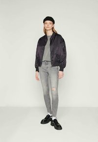 Young woman in black beret, black bomber jacket, gray t-shirt, ripped gray skinny jeans, and black platform shoes, standing against plain backdrop.