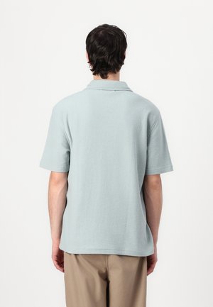 Homme aux cheveux foncés portant une chemise à manches courtes bleu clair et un pantalon beige, vu de dos sur un fond blanc uni.