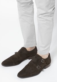 Donkerbruine suède dubbel-monk-schoenen met een spitse neus, decoratieve gespen en een lage gestapelde hak, gecombineerd met lichtgrijze taperende broek.