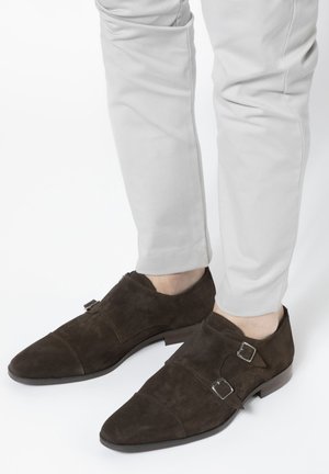 Chaussures à double boucle en suède marron foncé avec un bout pointu, des boucles décoratives et un petit talon empilé, associées à un pantalon fuselé gris clair.