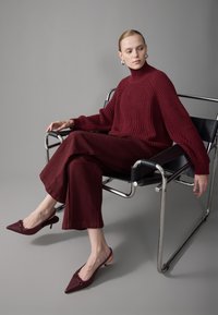 Bordeaux gestrickter Pullover, weite passende Hose und bordeauxfarbene Slingback-Absätze. Modell sitzt auf einem schwarzen Stuhl mit Chromakzenten.