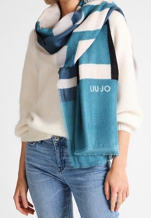 Femme portant un pull blanc et un jean bleu, avec une grande écharpe à rayures bleues et blanches drapée autour du cou et des épaules.
