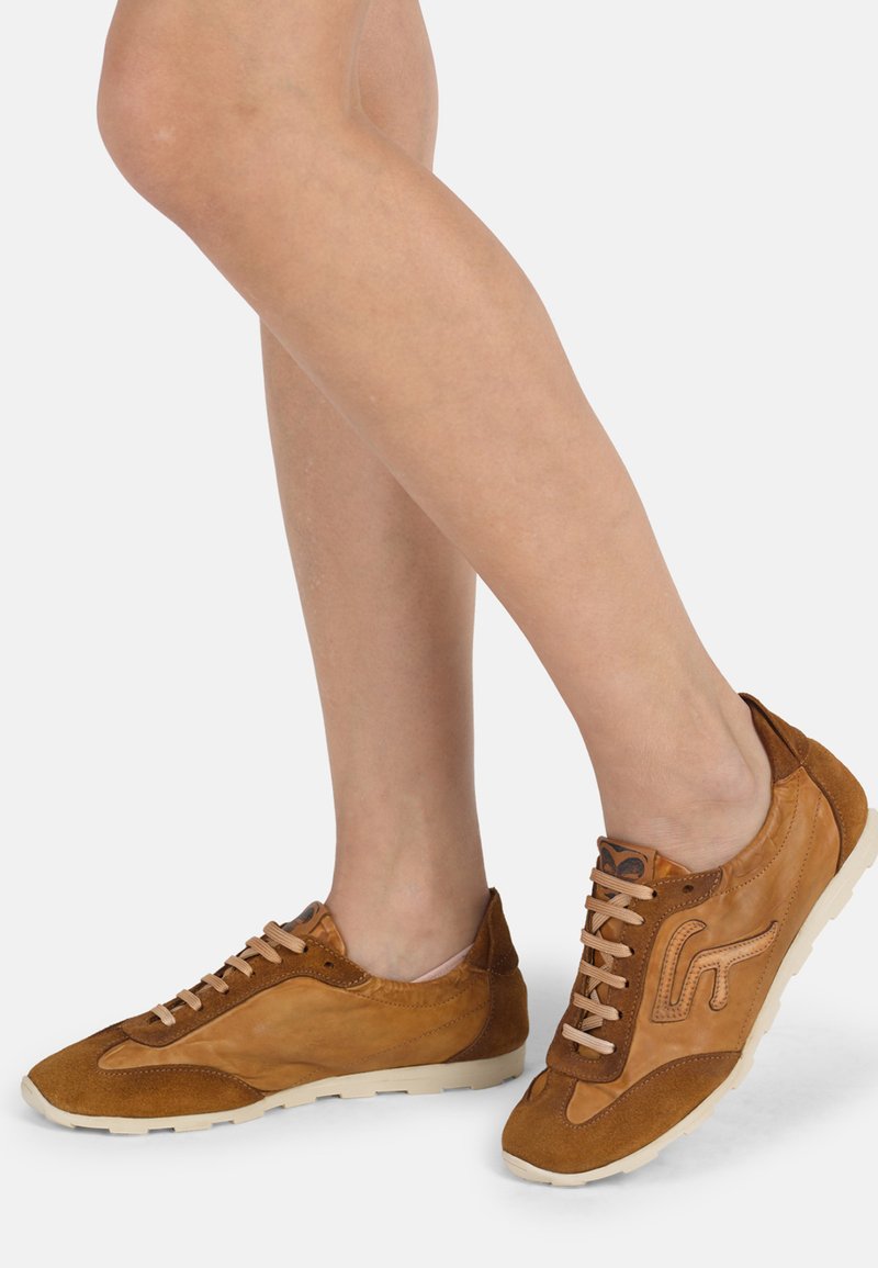 Jambes portant des baskets en cuir marron et en suède avec des semelles et des lacets beige, debout contre un fond clair uni.