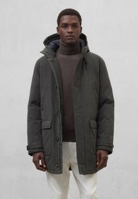 Ciemnozielona parka z zamkiem błyskawicznym i zapięciem na guziki, dwoma przednimi kieszeniami oraz kapturem. Noszona na brązowym swetrze z golfem i białych spodniach.