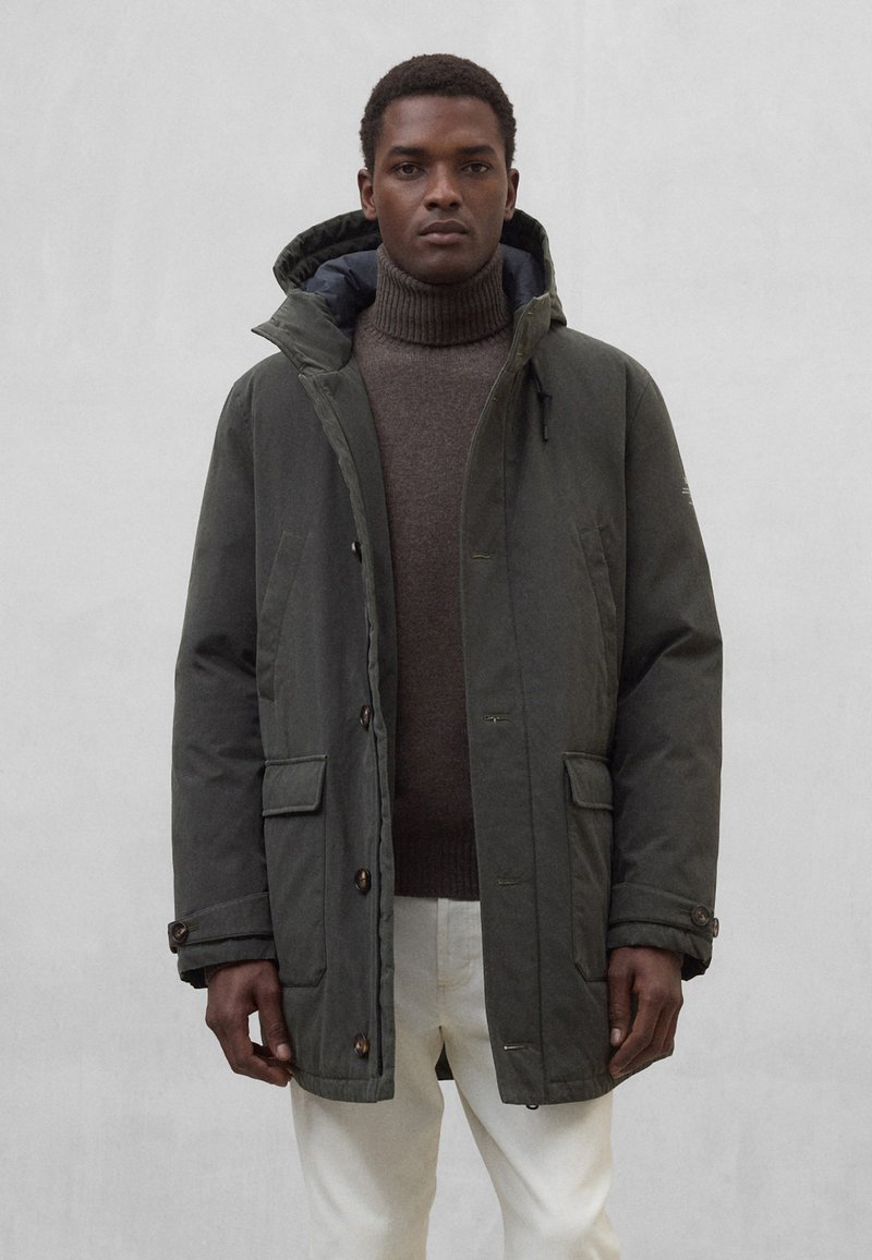 Ciemnozielona parka z zamkiem błyskawicznym i zapięciem na guziki, dwoma przednimi kieszeniami oraz kapturem. Noszona na brązowym swetrze z golfem i białych spodniach.