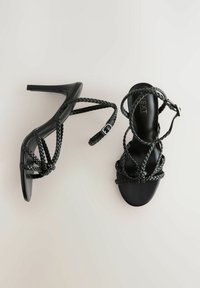 Schwarze High-Heels-Sandalen mit geflochtenen Riemen, offenes Zehendesign, glatte matte Textur und verstellbare Knöchelriemen für einen sicheren Sitz.