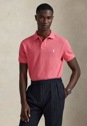 Homme portant un polo rose avec un logo blanc et un pantalon noir à fines rayures, debout avec une main dans la poche devant un fond neutre.