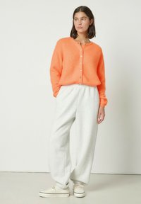 Oranžinis pūkuotas megztinis su sagomis, derinamas su šviesiai pilkomis sportinėmis kelnėmis. Audinio tekstūra yra minkšta, laisvo kirpimo. Avalynė – kreminės spalvos sportbačiai.