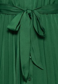 Grünes, plissiertes Stoffkleid mit einem breiten, passenden Gürtel, der in der Taille gebunden ist, und Knöpfen an der Vorderseite. Glatte Textur mit einer lebhaften Farbnuance.