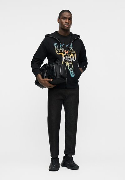 Άνδρας που φοράει μαύρο φούτερ με φερμουάρ και πολύχρωμο τύπωμα αστροναύτη, μαύρο παντελόνι και παπούτσια, κρατώντας μια μεγάλη μαύρη τσάντα στο ένα χέρι.