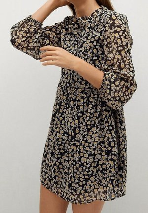 Femme portant une mini robe noire à imprimé floral avec des manches longues en voile transparent bouffantes et un col haut à volants, ajustant ses cheveux d'une main.