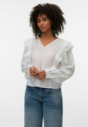 Göndör hajú nő, fehér fodros, hosszú ujjú blúzban és kék farmerral, egy egyszínű fehér háttér előtt áll.