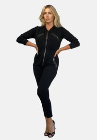 Czarny kombinezon z zamkiem z przodu, długimi rękawami oraz kieszeniami na zamek z przodu i po bokach. Dopasowane nogawki oraz elastyczny pas dla komfortu.