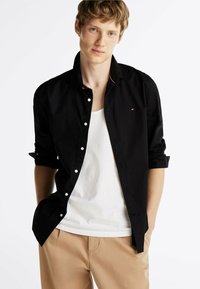 Chemise noire à manches longues avec une coupe décontractée, poignets à bouton, et un logo discret sur la poitrine. Portée par-dessus un débardeur blanc et un pantalon beige.